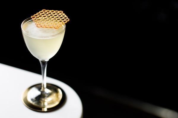 A glass of almost translucent drink with a honeycomb placed on the top, sits on a white surface. The background is black.
